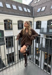 Brązowy kardigan z prążkowanej dzianiny z dużymi guzikami, w połączeniu z brązowymi szortami z białymi paskami oraz brązowymi botkami do kolan. Tło z metalowym balustradą.