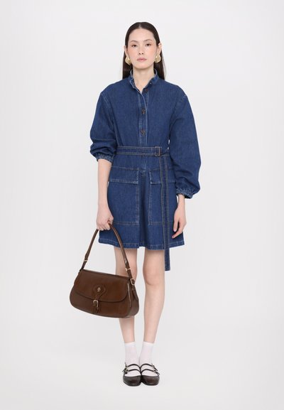 Vestido de camisa de mezclilla con mangas largas, cintura ceñida y dos bolsillos frontales. Combinado con un bolso de cuero marrón y calcetines blancos hasta el tobillo.