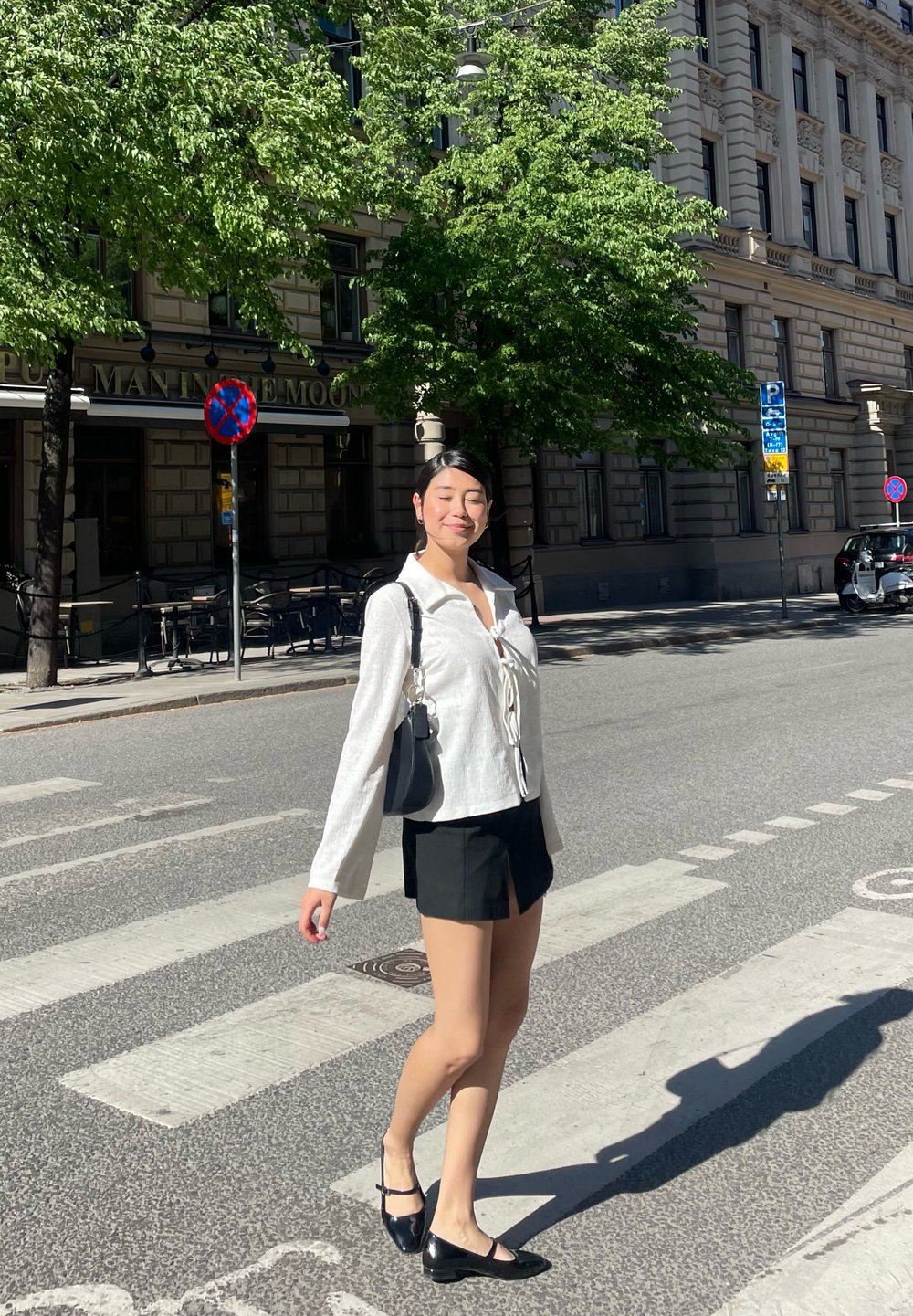 Λευκό πουκάμισο με μακριά μανίκια και σχέδιο γραβάτας, συνδυασμένο με μια μαύρη μίνι φούστα. Μαύρη τσάντα και παπούτσια, standing σε διάβαση πεζών κοντά σε πράσινο χώρο.