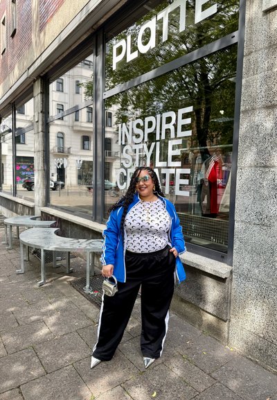 Chaqueta azul con cremallera, camiseta blanca con patrones de luna creciente, pantalones negros con franjas blancas, zapatos plateados de punta y sosteniendo un bolso metálico.
