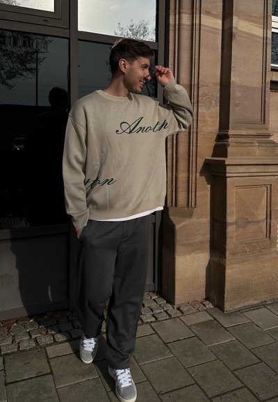 Suéter de punto beige con texto verde, pantalones grises y zapatillas gris claro. Modelo de pie afuera, ajustándose el cabello cerca de un edificio de piedra.