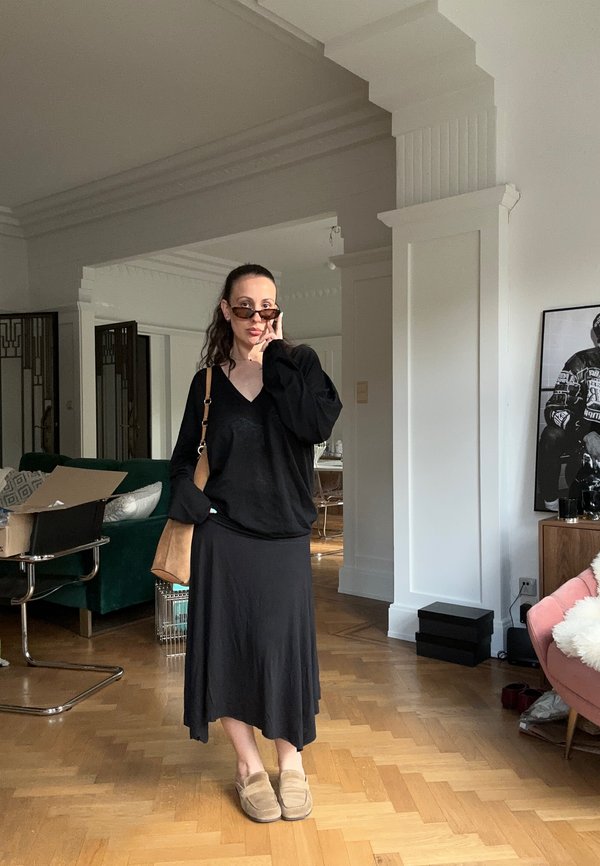 Femeie cu păr șaten, ochelari de soare, top negru, fustă neagră, geantă bej și papuci, stă într-un living elegant cu podea din parchet.