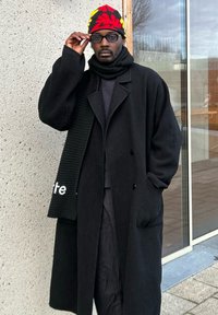 Casaco de trench em lã, na cor preta e tamanho grande, sobre um suéter texturizado preto, complementado por um gorro de malha colorido e um cachecol preto canelado.