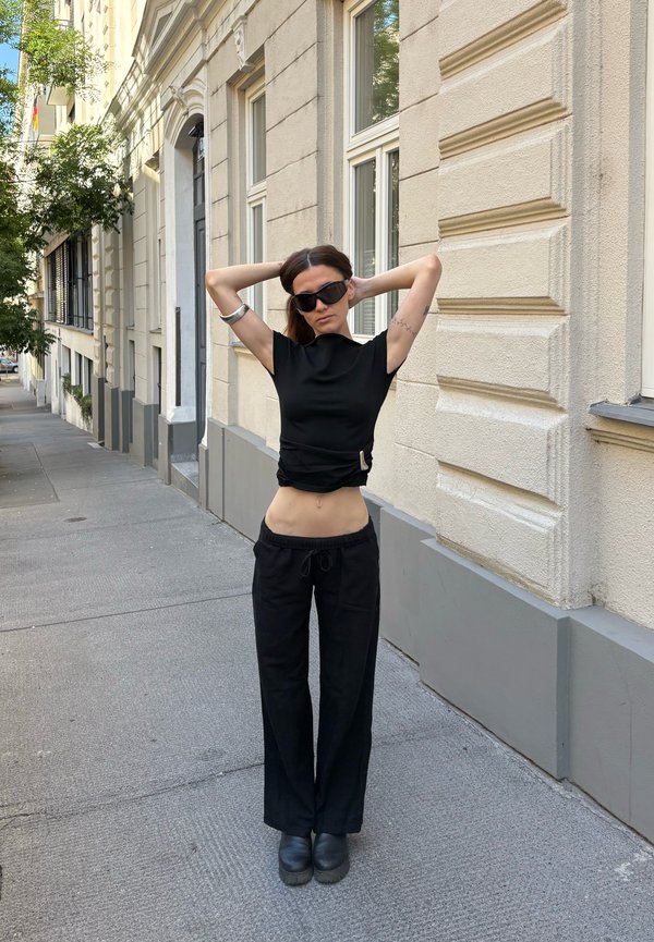 Femme en tenue noire et lunettes de soleil debout sur un trottoir de la ville, les mains derrière la tête, près d’un bâtiment beige avec fenêtres.