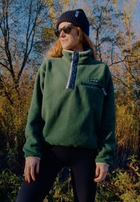 Zielony pulower z polaru z wysokim kołnierzem, niebieską plisą na guziki, kieszenią na piersi na zamek oraz kontrastującymi białymi mankietami. Czarne okulary przeciwsłoneczne i czapka typu beanie.