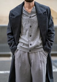 Casaco cinza escuro sobre um suéter claro de botão cinza, combinado com calças cinza pregueadas. Tecido texturizado com cores contrastantes.