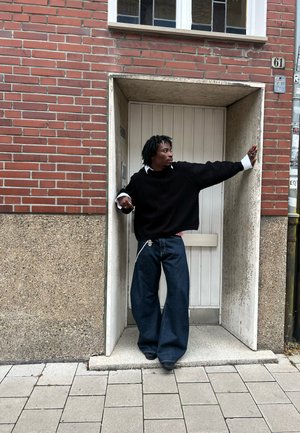 Jersey negro de gran tamaño, camisa blanca debajo y vaqueros de pierna ancha de mezclilla oscura. De pie en un umbral con un fondo de ladrillo y piedra.