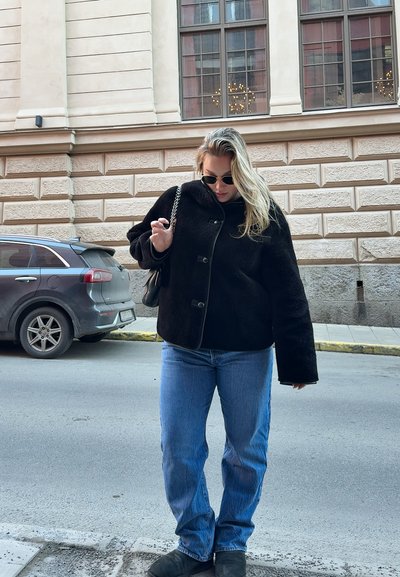 Chaqueta corta negra de felpa con grandes botones y cuello alto; combinada con unos vaqueros de un azul claro y calzado negro, de pie en una calle de la ciudad.