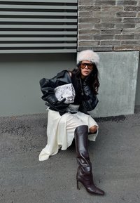 Schwarze Lederjacke über einem schwarzen Top, weißer Rock mit Spitzenbesatz, braune strukturierte Overkneestiefel und ein hellrosa Pelzhut.