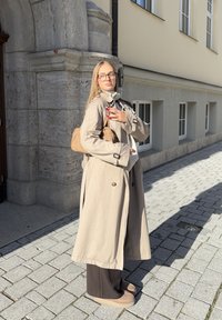 Žena v béžovém trenčkotě, hnědých kalhotách a brýlích, nosící světle hnědou kabelku, stojí na sluncem osvětlené dlažební ulici poblíž kamenné budovy.