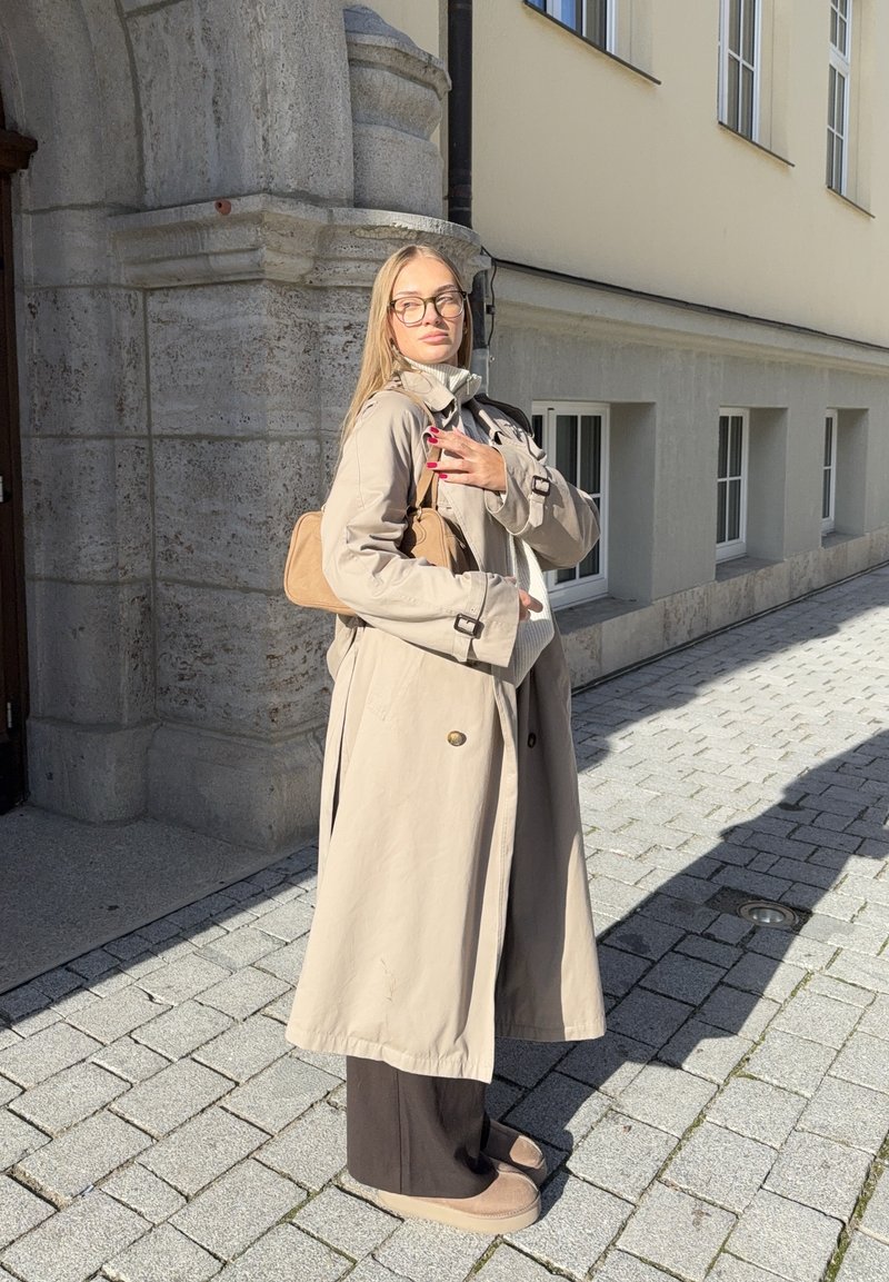 Kvinde i beige trenchcoat, brune bukser og briller, der bærer en lys taske, står på en solbeskinnet brostensbelagt gade nær en stenvæg.