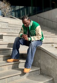 Άνδρας με πλεξούδες που φοράει πράσινο varsity μπουφάν, γραβάτα, τζιν και καφέ μποτάκια, καθισμένος σε εξωτερικές σκάλες από σκυρόδεμα, κοιτάζοντας τα χέρια του.