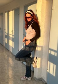 Lyserød bluse i blonder over sort langærmet skjorte, blå jeans og grå sneakers med lyserøde detaljer; lilla solbriller; står op ad en væg.