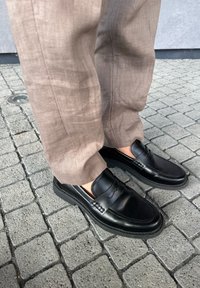 Crni kožni loafers s glatkom površinom, s dekorativnim šavovima i debelim gumiranim potplatom. U kombinaciji s bež lanenim hlačama.