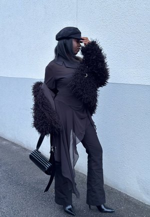 Mujer con vestido negro de mangas esponjosas y gorra sostiene un bolso negro, de pie frente a una pared texturizada blanca y gris.
