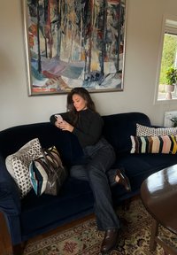 Kobieta w czarnej bluzce i dżinsach siedzi na granatowej kanapie, korzystając ze smartfona, w otoczeniu wzorzystych poduszek, toreb, abstrakcyjnego obrazu, okna i roślin.