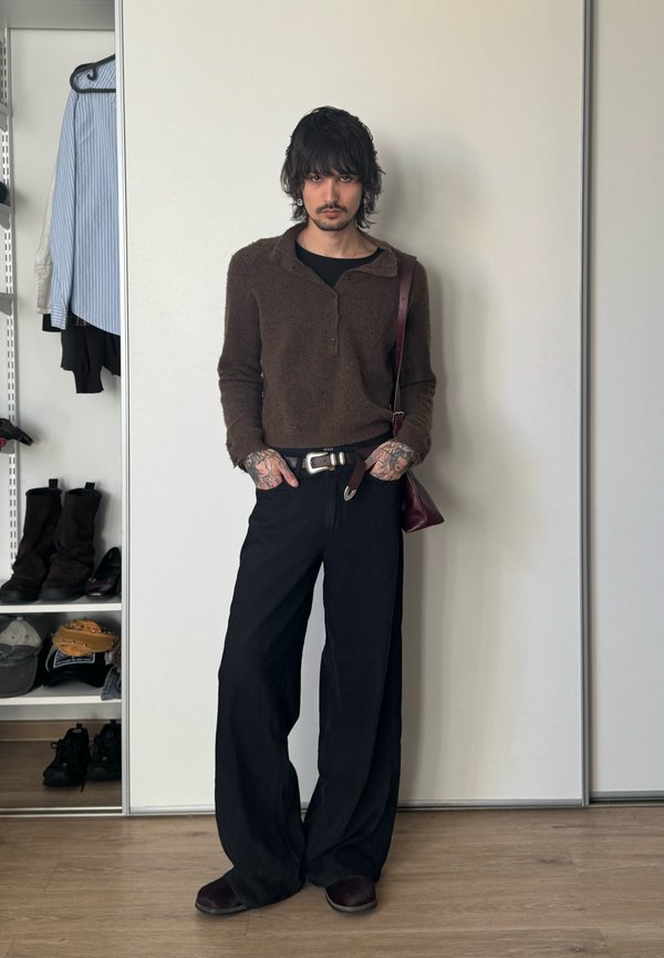 Homme aux cheveux foncés et tatouages portant un pull marron, un pantalon large noir et un sac à bandoulière bordeaux, debout à l'intérieur.