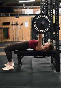 Des poids de barre sur un banc de musculation. La personne porte un haut de sport bordeaux et des leggings foncés, avec des baskets claires. Sol en caoutchouc noir.