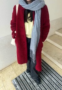 Cappotto rosso e soffice con finitura testurizzata, abbinato a un maglione giallo chiaro e una sciarpa grigia. Scarpe nere e jeans scuri completano il look.