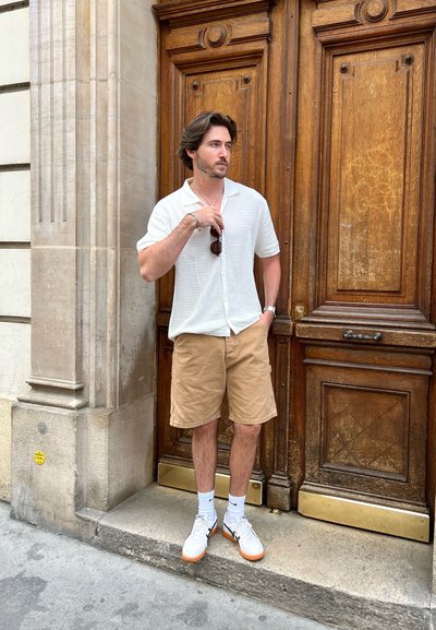 Camisa blanca de manga corta con textura, pantalones cortos beige, zapatillas blancas con suelas naranjas. De pie frente a una puerta de madera con paneles decorativos.