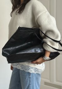 Sac en cuir noir avec une surface texturée, une forme angulaire et deux poignées, tenu contre un pull de couleur claire et un ourlet en dentelle.