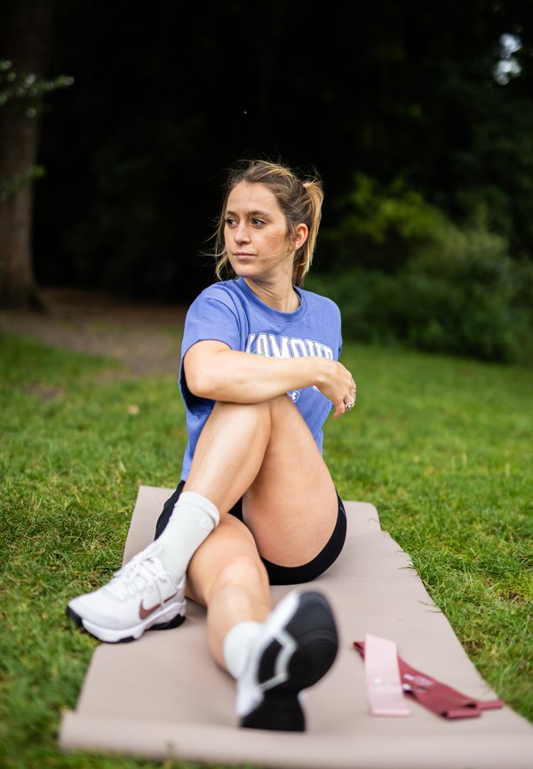 Camiseta azul con el texto "Komor", pantalones cortos negros, zapatos Nike blancos. Sentado sobre un tapete beige en la hierba, con dos bandas de resistencia en rosa y burdeos.
