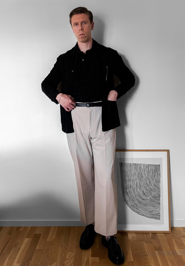 Mann in schwarzer Jacke und Hemd mit beigen Hosen und schwarzen Schuhen steht neben einem gerahmten schwarz-weißen abstrakten Linienkunstwerk auf Holzboden.