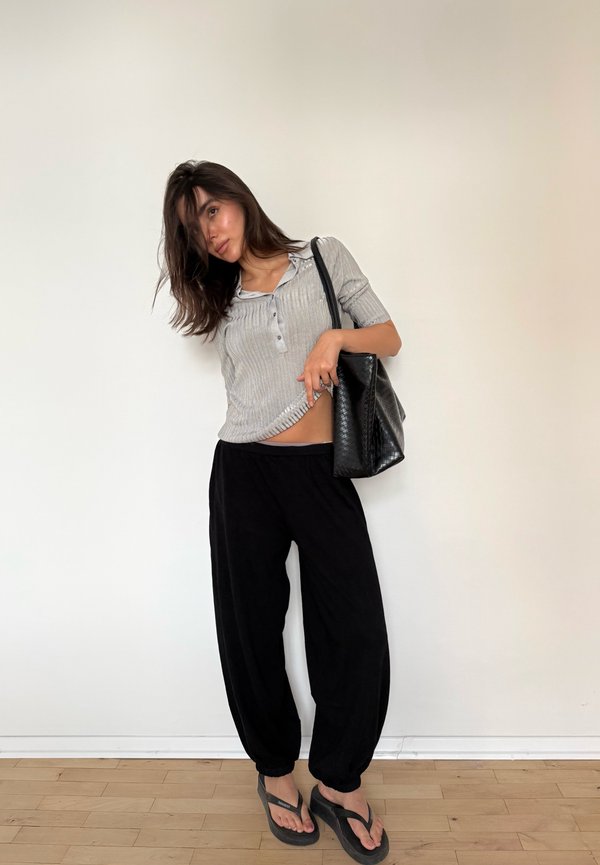 Woman in gray short-sleeve top and black pants holding a black handbag, standing with one hand lifting her shirt slightly against a plain wall.