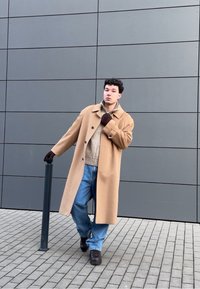 Manteau en laine beige sur un pull marron clair, jeans bleu amples, chaussures noires et gants marron. Debout à côté d'un poteau sombre sur une surface carrelée.