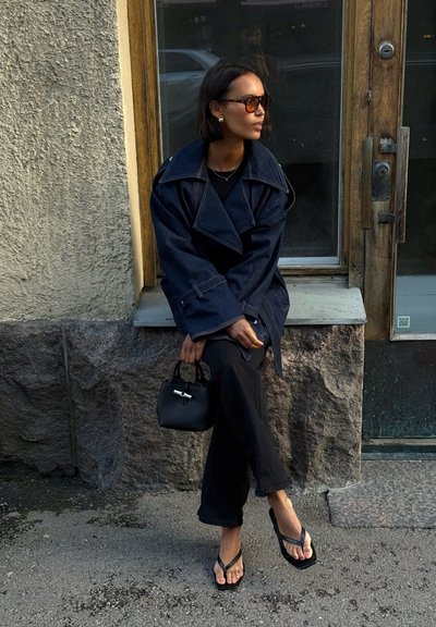Trench coat de denim oscuro, blusa negra, pantalones relajados negros, chanclas negras, sosteniendo un pequeño bolso negro y con unas gafas de sol marrones de gran tamaño.