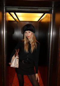 Jeune femme aux longs cheveux blonds, vêtue d'une tenue noire et d'un chapeau en fourrure, tenant un sac beige, debout dans un ascenseur aux parois en bois faiblement éclairé.