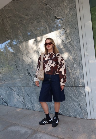 Joven mujer con gafas de sol, chaqueta de estampado de vaca, pantalones cortos de mezclilla, zapatillas negras de Adidas y llevando un bolso de hombro beige, de pie contra una pared de piedra.