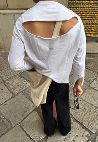 Chemise blanche à manches longues avec des découpes dans le dos, portée avec un pantalon large noir. Un sac fourre-tout beige pâle pend d'un côté, contenant des lunettes de soleil.