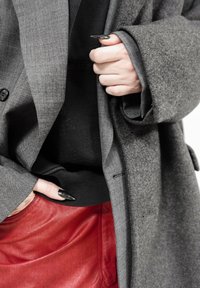 Gros plan d'une personne portant des manteaux gris superposés, un pull noir et une jupe en cuir rouge, avec des ongles longs noirs pailletés visibles sur la main.