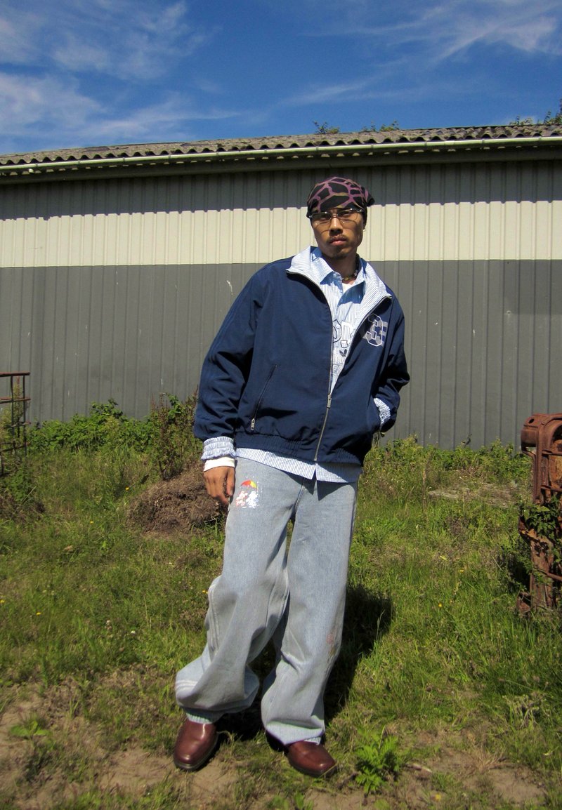 Jeune personne debout en plein air sur l'herbe portant une veste bleu marine, un jean large bleu clair avec un patch, un foulard à motifs et des chaussures marron.