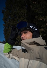 Skiová bunda v béžovej a zelenej farbe, detailný záber muža, ktorý si upravuje čiernu prilbu so zrkadlovými okuliarmi v zasneženom prostredí. Čierne stromy v pozadí.