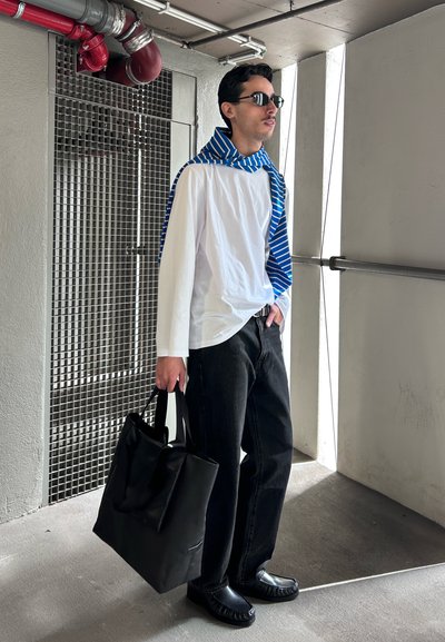 Hombre con gafas de sol, camisa blanca de manga larga con bufanda de rayas azules, pantalones negros, sosteniendo un bolso negro, de pie en un pasillo industrial.