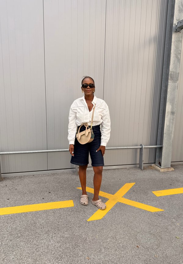 Frau mit weissem Hemd, dunklen Shorts, Sonnenbrille und beigen Crocs steht auf grauem Pflaster mit gelben Straßenmarkierungen neben einer grauen Metallwand.