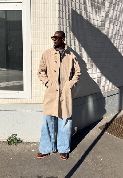 Impermable beige con botones, pantalones de mezclilla anchos de tiro alto en un tono azul claro y zapatos de plataforma marrones. Gafas de sol de gran tamaño completan el look.