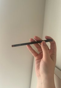 Matita per labbra MAC tenuta tra le dita, con un corpo nero e una punta rosa scura, texture liscia, con un singolo dettaglio del logo in argento.