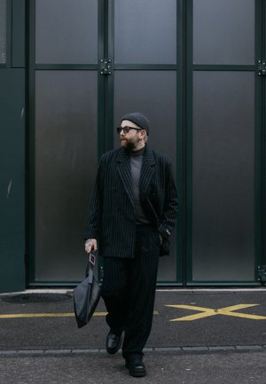 Traje negro a rayas con abrigo ajustado y pantalones anchos. Suéter de cuello alto gris, gorro negro, gafas de sol cuadradas y llevando una bolsa negra.
