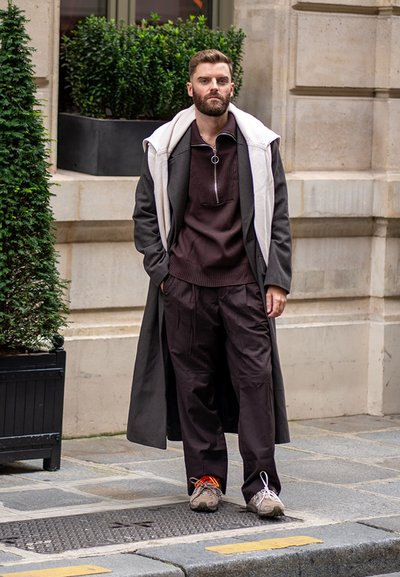 Largo abrigo negro sobre un suéter de punto marrón oscuro con cuello con cremallera, combinado con pantalones holgados oscuros y zapatillas deportivas con cordones en contraste.