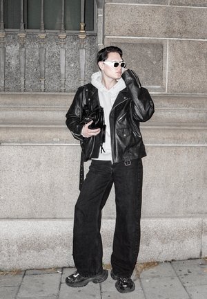 Hombre con gafas de sol blancas, chaqueta de cuero negra, sudadera gris, pantalones anchos negros y zapatillas, de pie junto a una pared de piedra sosteniendo una bolsa negra.