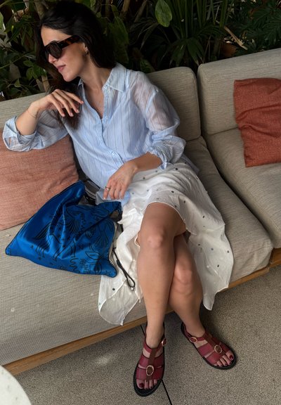 Mujer con gafas de sol, camisa azul claro a rayas, falda blanca y sandalias rojas sentada en un sofá beige con cojines naranjas y bolsa azul cerca.