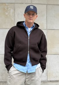 A man wearing a black zip-up hoodie over a blue button-up shirt, beige trousers, round glasses, and a navy baseball cap is standing against a tiled wall.