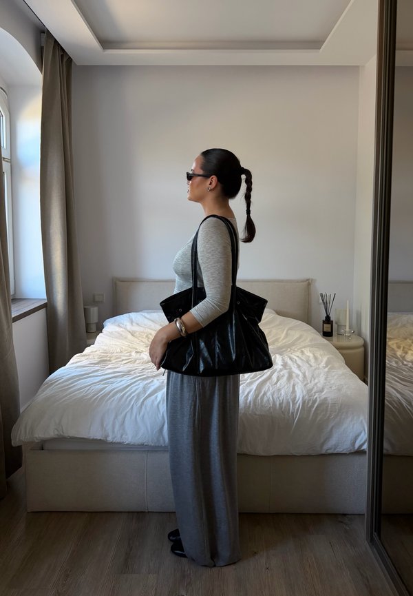 Una mujer con gafas de sol y un vestido largo gris está de pie junto a una cama y lleva un bolso grande negro al hombro.