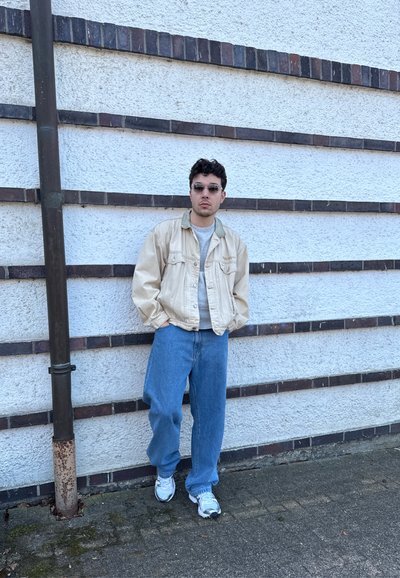 Joven con gafas de sol, chaqueta beige, camisa gris, vaqueros azules holgados y zapatillas blancas, de pie frente a una pared blanca texturizada con franjas horizontales oscuras.