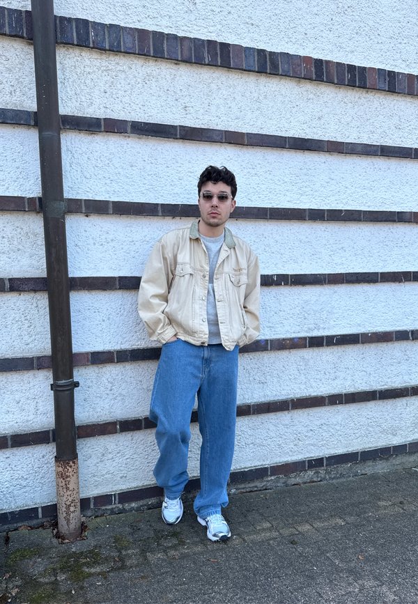 Νέος άνδρας φορώντας γυαλιά ηλίου, μπεζ μπουφάν, γκρι πουκάμισο, φαρδιά μπλε τζιν παντελόνια και λευκά αθλητικά παπούτσια στέκεται μπροστά σε μια υφή λευκού τοίχου με οριζόντιες σκοτεινές ρίγες.