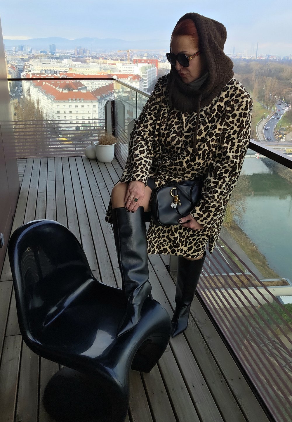 Chaise noire brillante, manteau imprimé léopard, bottes en cuir noires montant jusqu'aux genoux, et sac à main noir. Balcon urbain avec panorama de la ville en arrière-plan.