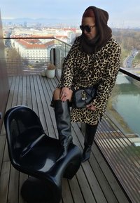 Chaise noire brillante, manteau imprimé léopard, bottes en cuir noires montant jusqu'aux genoux, et sac à main noir. Balcon urbain avec panorama de la ville en arrière-plan.